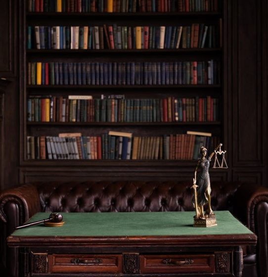 A classic study room with shelves of books, a gavel, and Lady Justice figurine on a green table.
